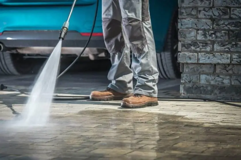 A person is pressure washing a paved surface near a blue car, focusing on the ground near their feet.