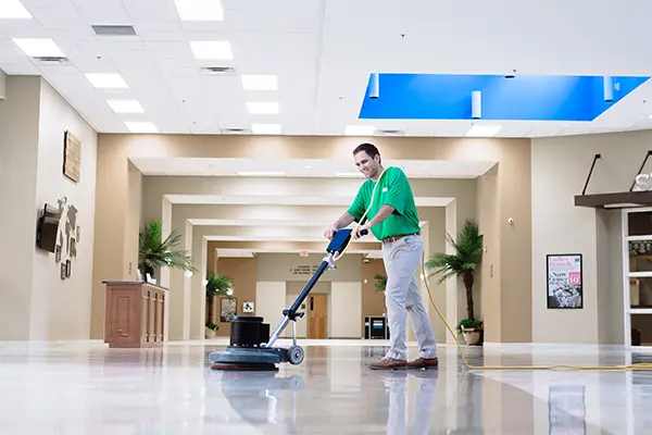 A person is using an industrial floor cleaning machine inside a spacious warehouse with tall shelving stocked with boxes.