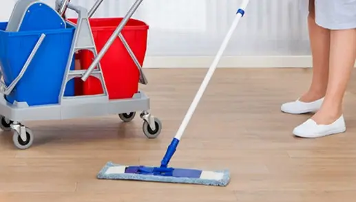 A person in white shoes is mopping a floor next to a blue and red janitorial cart.