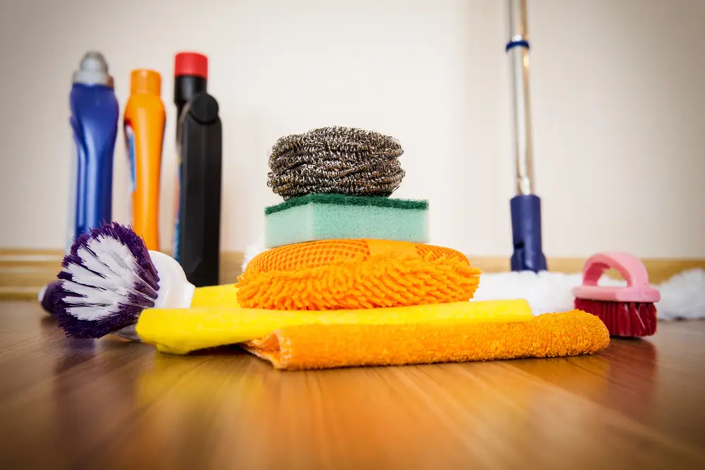 Various cleaning supplies including sponges, brushes, a mop, and detergents on a wooden floor against a wall.