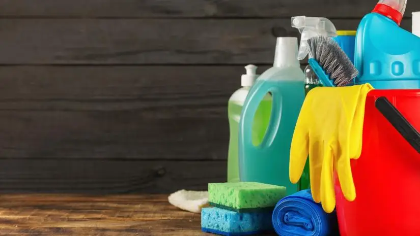 Various cleaning supplies, including detergents, gloves, brushes, and sponges, are arranged against a wooden background.